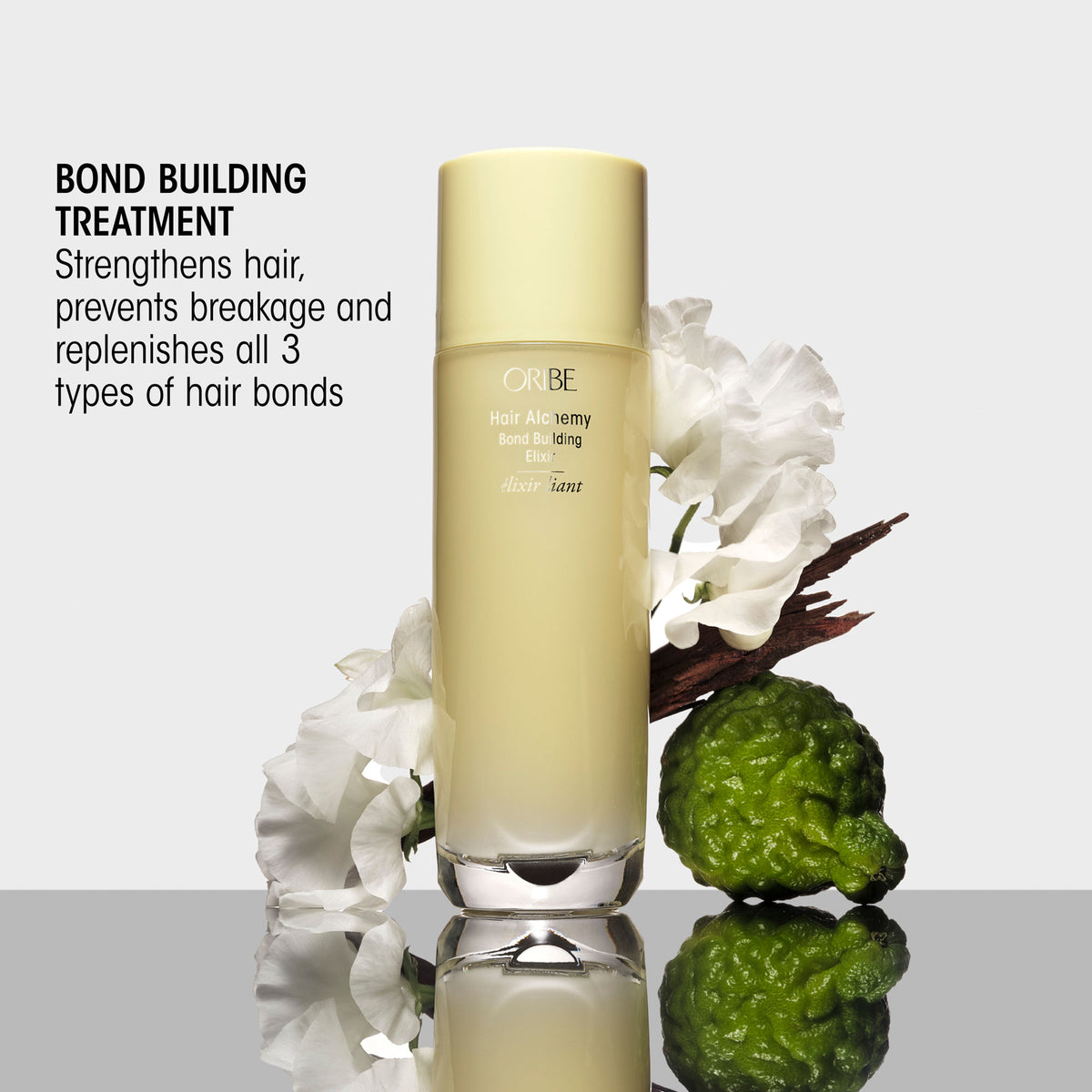 An Oribe Hair Alchemy Bond Repair Leave-In Treatment bottle stands next to white flowers and a green citrus fruit, formulated to strengthen hair, prevent breakage, and replenish all three types of hair bonds.