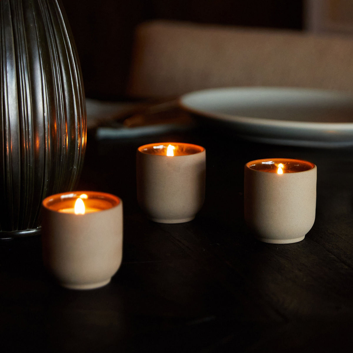 Three small, lit scented votives from the Homecourt Candle Discovery Set (Limited Edition) in matte tan ceramic vessels placed on a dark table.