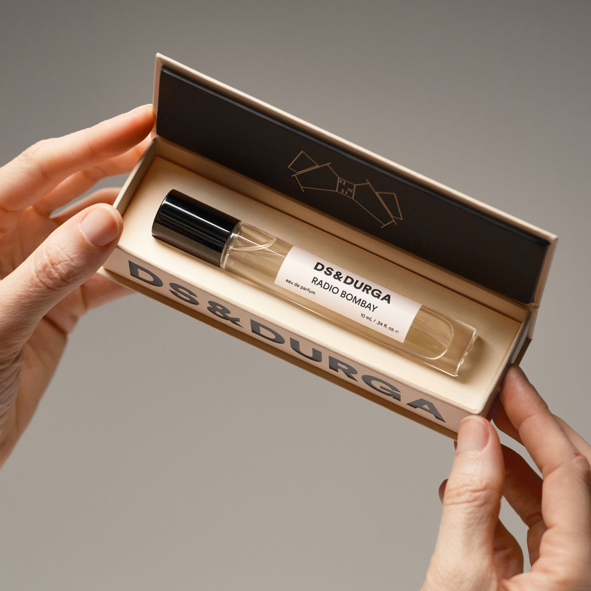 Hands hold an open cardboard box containing a cylindrical glass perfume rollerball of D.S. & Durga Radio Bombay Eau de Parfum, its scent reminiscent of warm Bandra ragas.