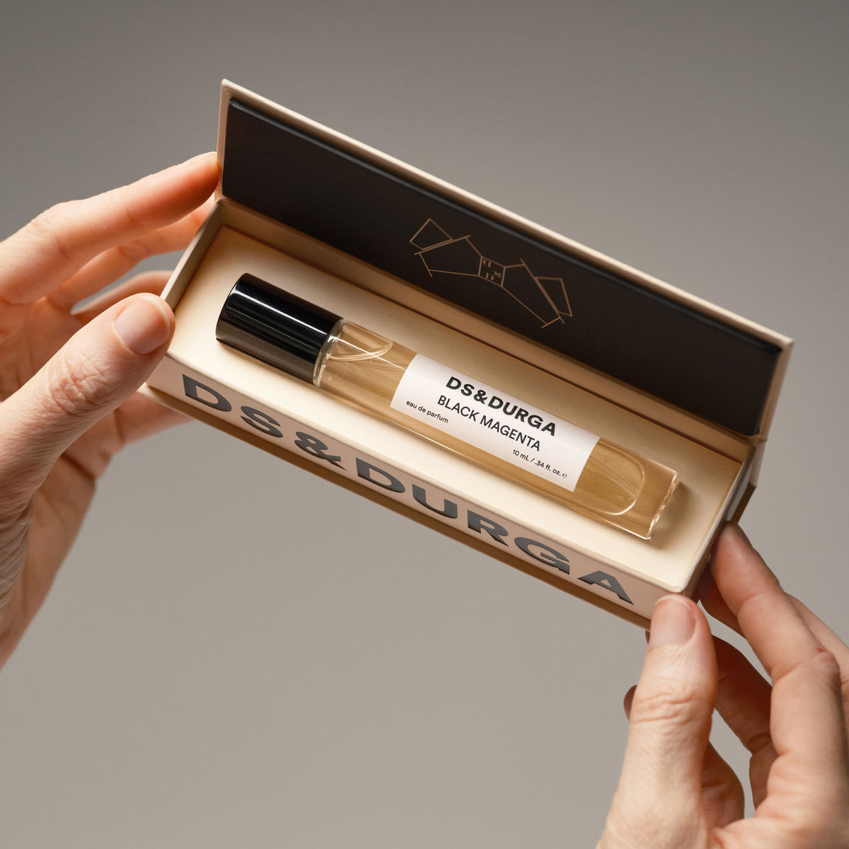 Hands holding an open rectangular box containing a Black Magenta Eau de Parfum glass rollerball vial with a black amber aesthetic.