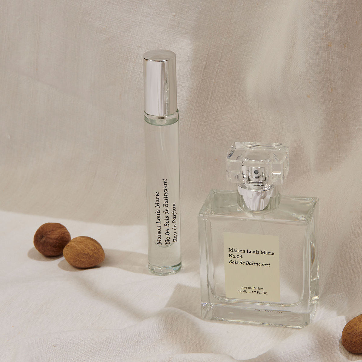 A cylindrical rollerball and a square glass bottle of Maison Louis Marie No.04 Bois de Balincourt Eau de Parfum rest on draped fabric next to three whole nutmegs, evoking fresh forests and the smell after rain.