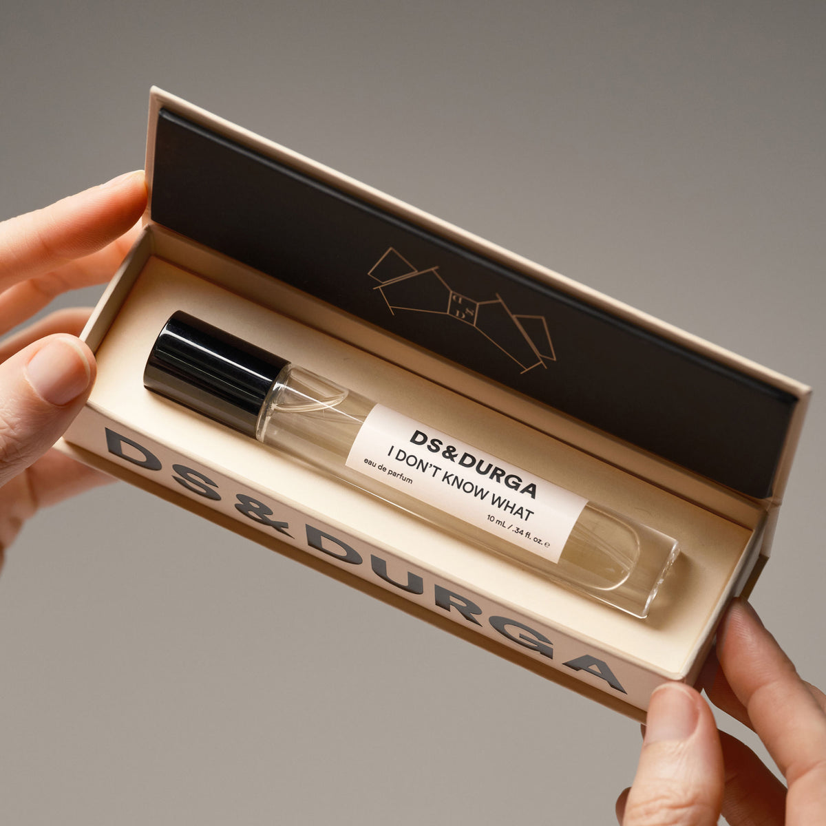 Hands holding an open D.S. & Durga I Don’t Know What Eau de Parfum box containing a glass rollerball vial, a fragrance enhancer for transparent radiance.