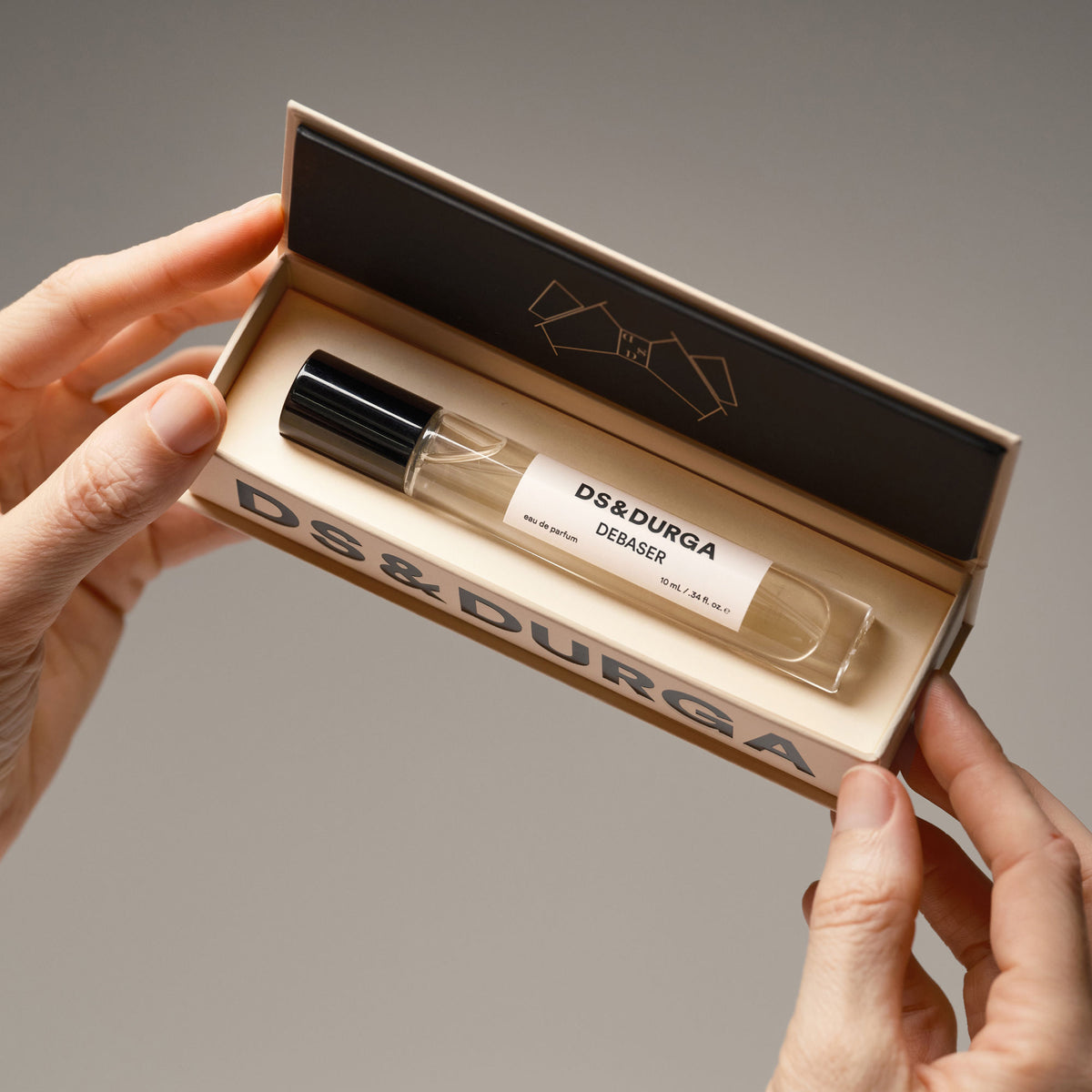 Hands hold open a small, rectangular box containing a glass rollerball vial of Debaser Eau de Parfum, evoking the atmosphere of August heat.