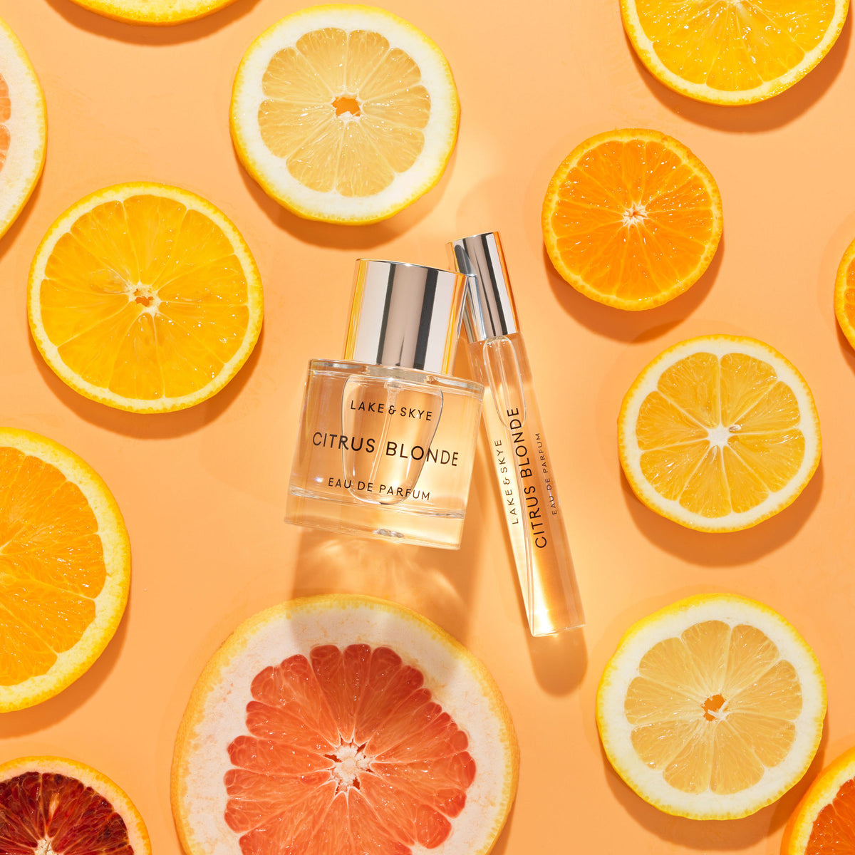 A full-size bottle of Lake & Skye Citrus Blonde Eau de Parfum and a travel spray vial surrounded by assorted citrus slices on an orange background.