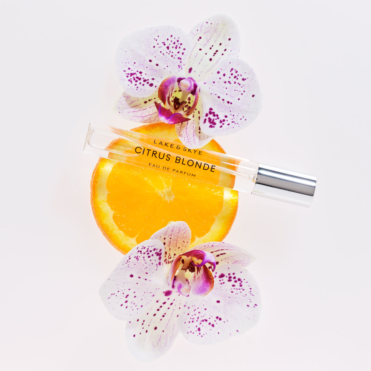 A Lake & Skye Citrus Blonde Eau de Parfum glass rollerball with a silver cap rests on a sliced orange between two white orchid flowers.