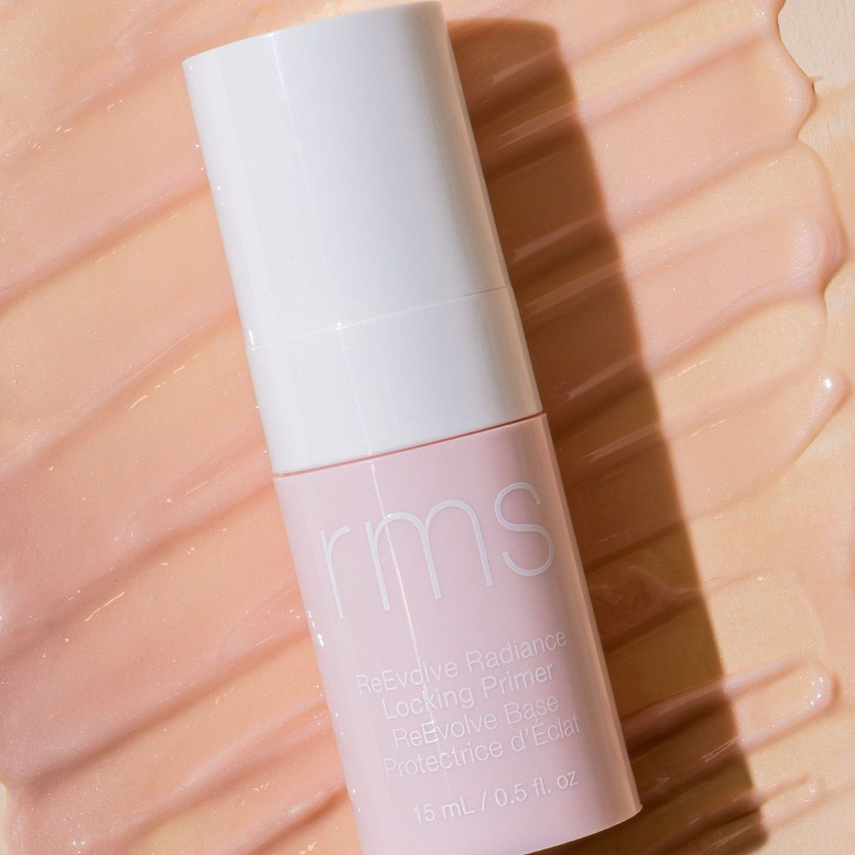A cylindrical refillable pump bottle of RMS Beauty ReEvolve Radiance Locking Primer resting on a smear of light-colored gel.