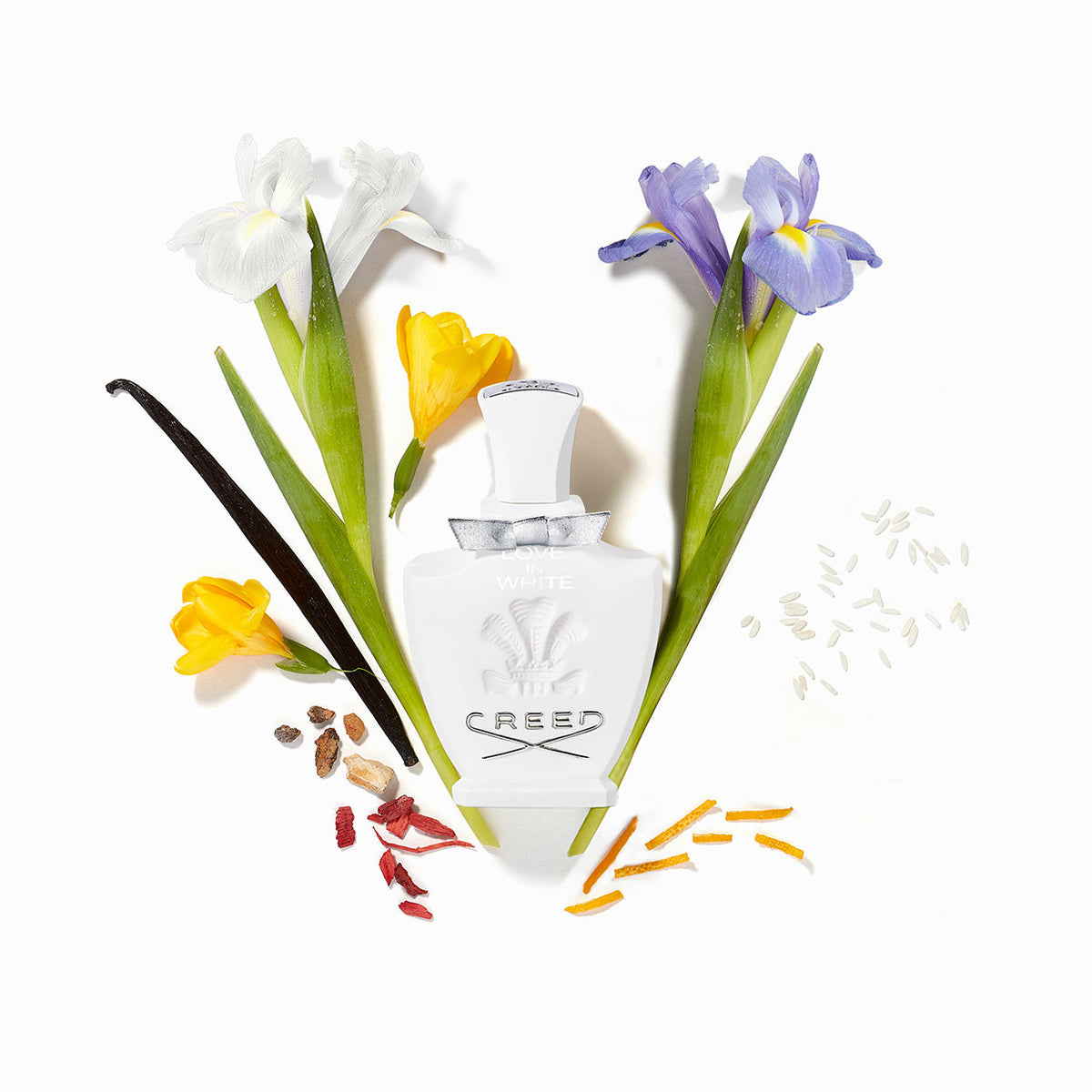 A white Creed Love In White Eau de Parfum bottle surrounded by iris flowers, a vanilla bean, grains of rice, and citrus peel.