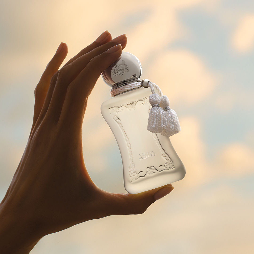 A hand holds a frosted glass Valaya Exclusif Parfum bottle with a silver cap and a white tassel against a sunset sky, embodying feminine elegance and refined luxury fragrance.