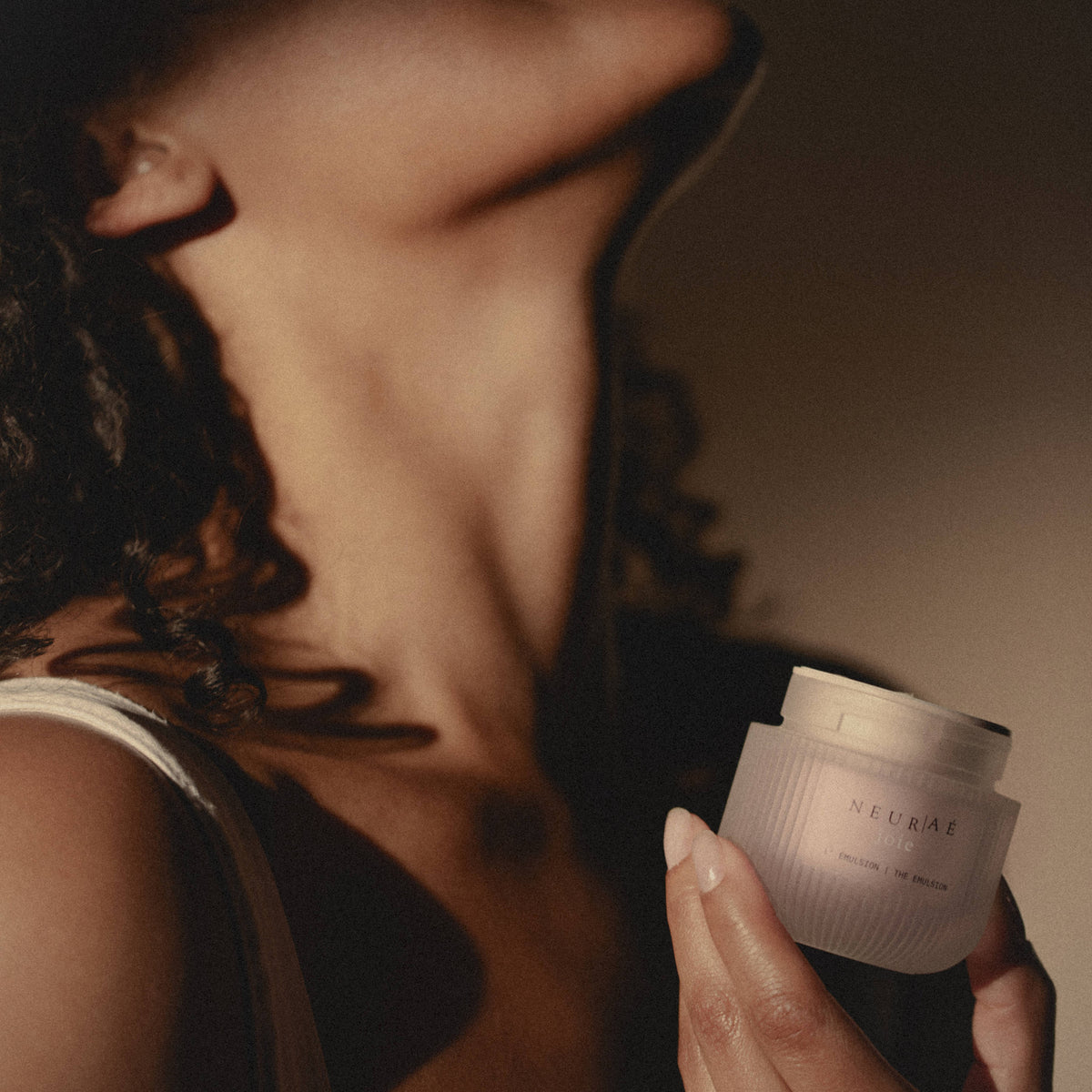 A person holds a small, ribbed glass jar of Neuraé Joie The Emulsion radiance cream.