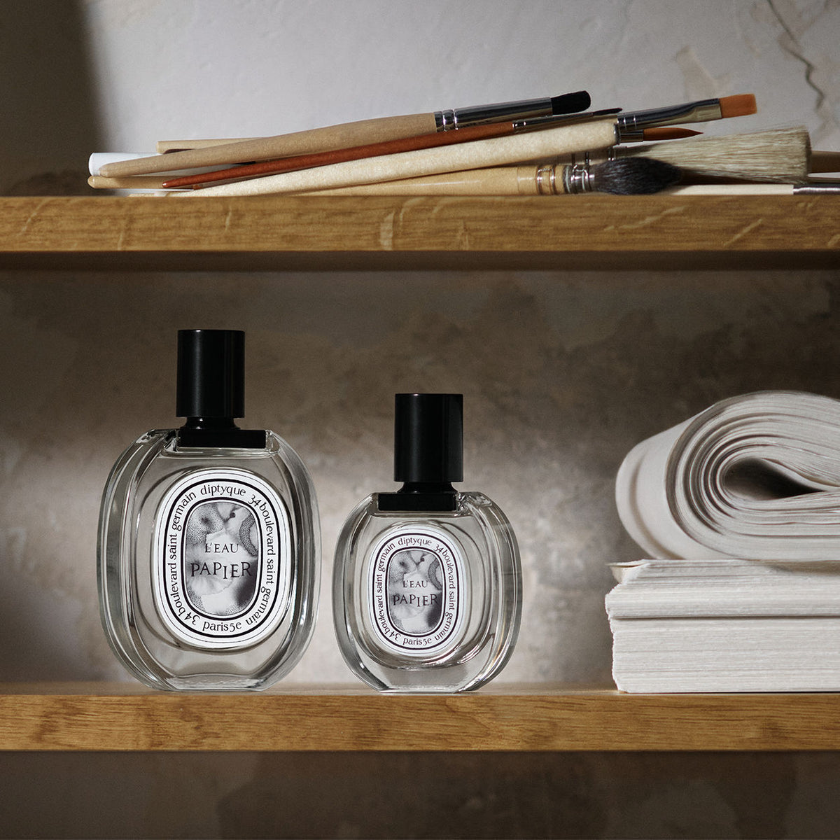 Two glass perfume bottles of different sizes sit on a wooden shelf next to a stack of L’Eau Papier Eau de Toilette, with paintbrushes resting on the shelf above.