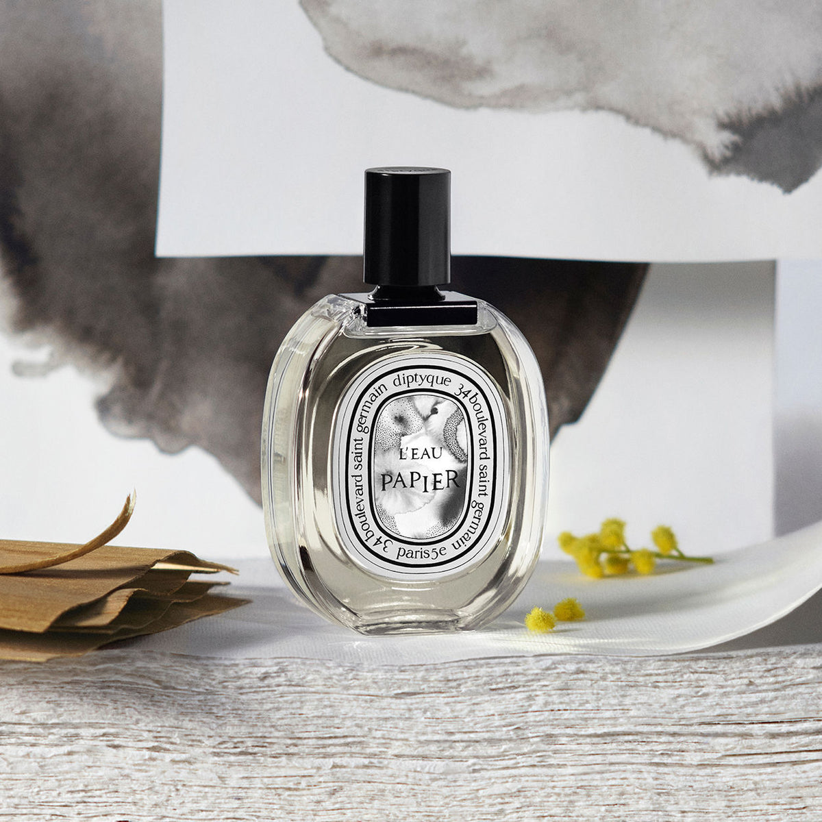 L’Eau Papier Eau de Toilette in an oval glass bottle with a black cap and illustrated label, resting on a textured surface with small yellow flowers.