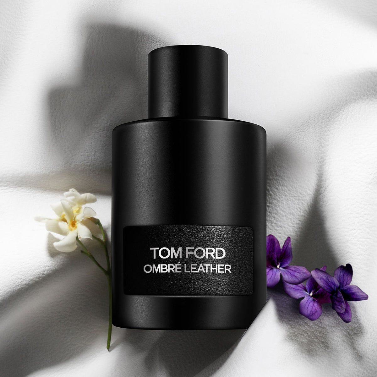 A matte black bottle of Tom Ford Ombre Leather Eau de Parfum with a textured label, flanked by a small white flower and purple blossoms on draped white fabric.