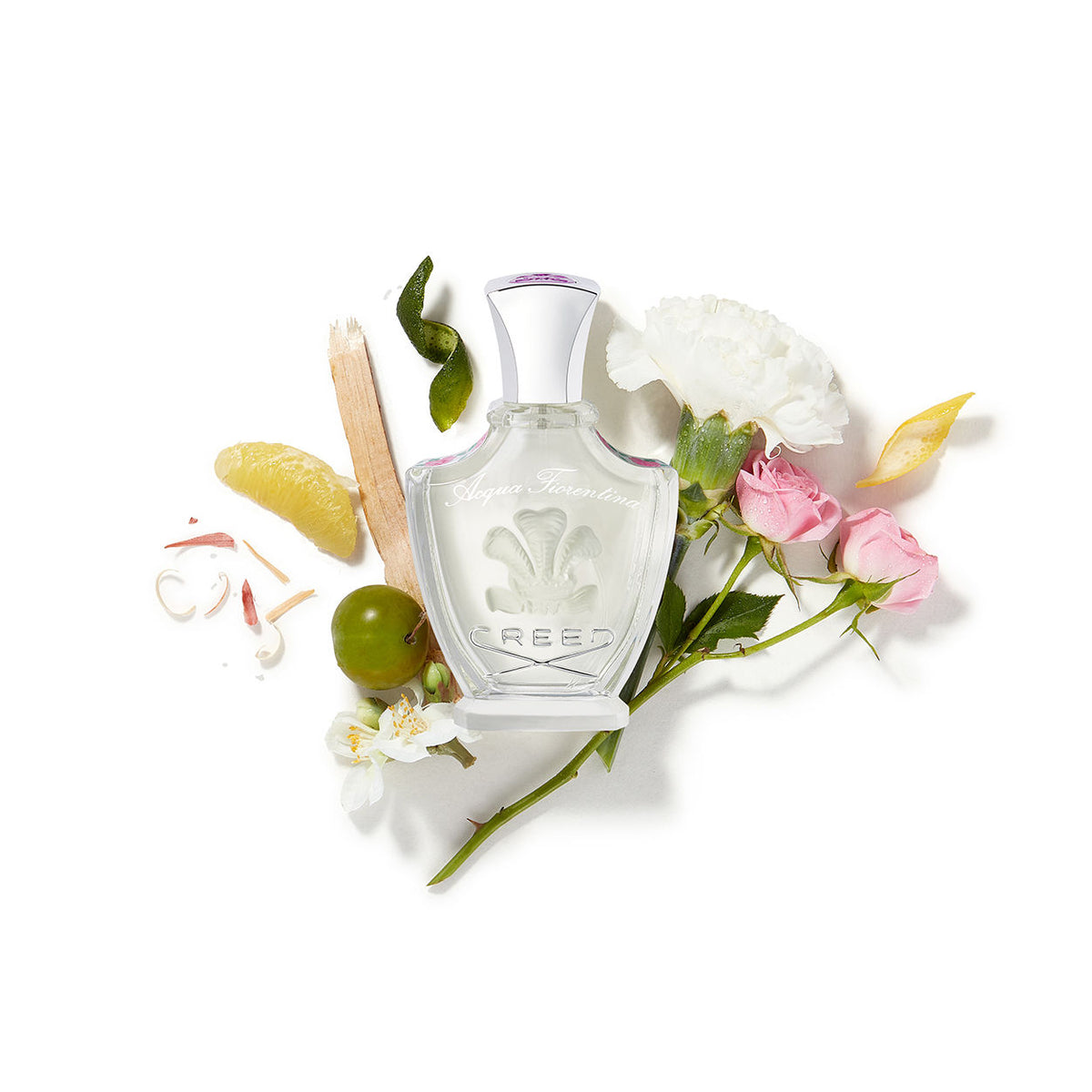 A bottle of Creed Acqua Fiorentina Eau de Parfum surrounded by a white carnation, two pink roses, a citrus slice, and small green fruit against a white background.