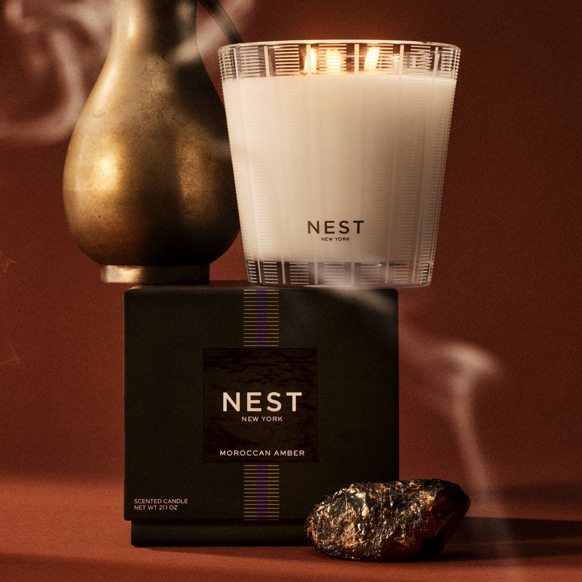 A Nest New York Moroccan Amber Candle in a glass jar sits atop its black box, next to a metallic vessel and a piece of raw amber.
