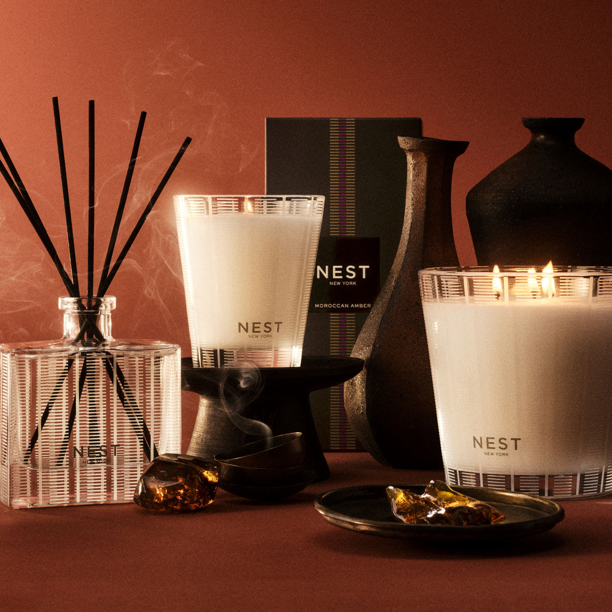 Two classic Moroccan Amber Candle fragranced candles with multiple wicks, a reed diffuser, and a dark ceramic vase arranged with amber pieces on a reddish-brown surface.