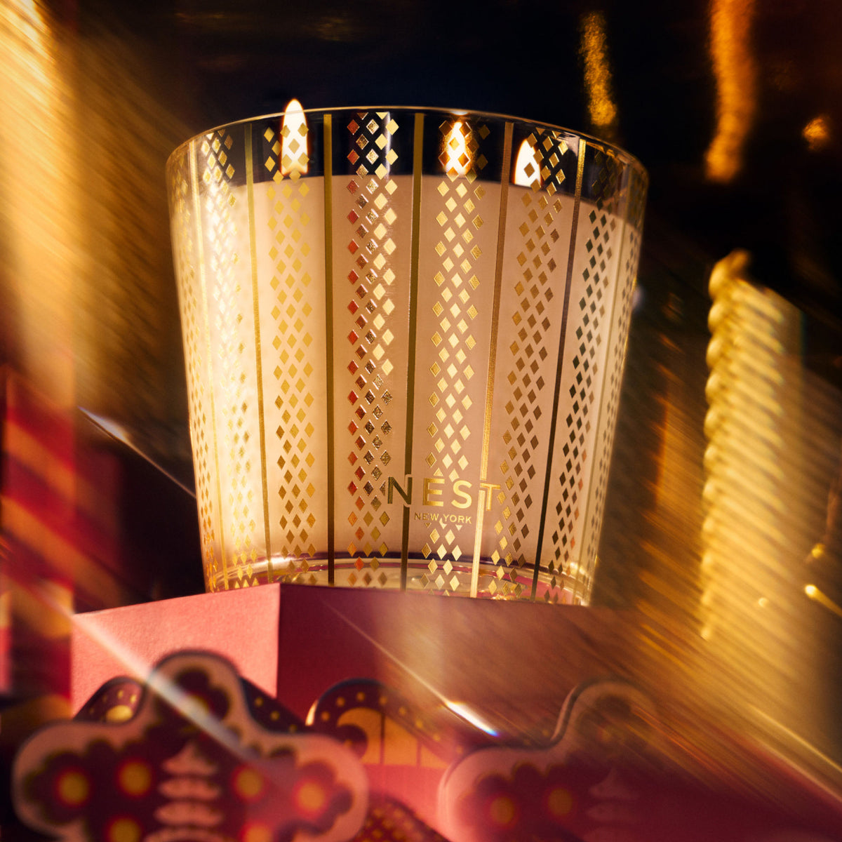 A three-wick Nest New York Holiday Candle (Limited Edition) in a glass vessel with a gold diamond pattern, glowing with a seasonal fragrance.