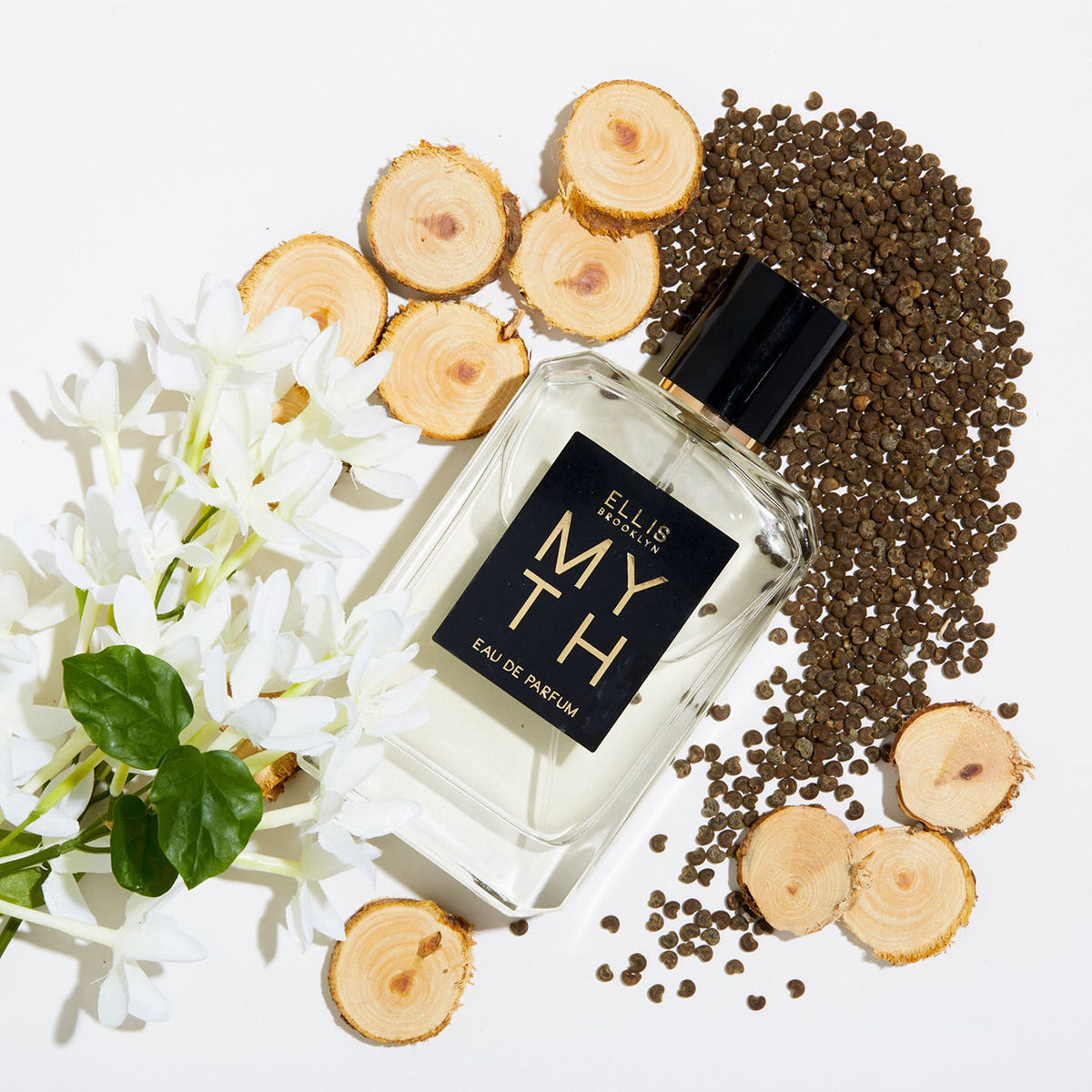 A rectangular MYTH Eau de Parfum glass bottle with a black label surrounded by jasmine petals, white cedarwood slices, and scattered seeds on a white surface.