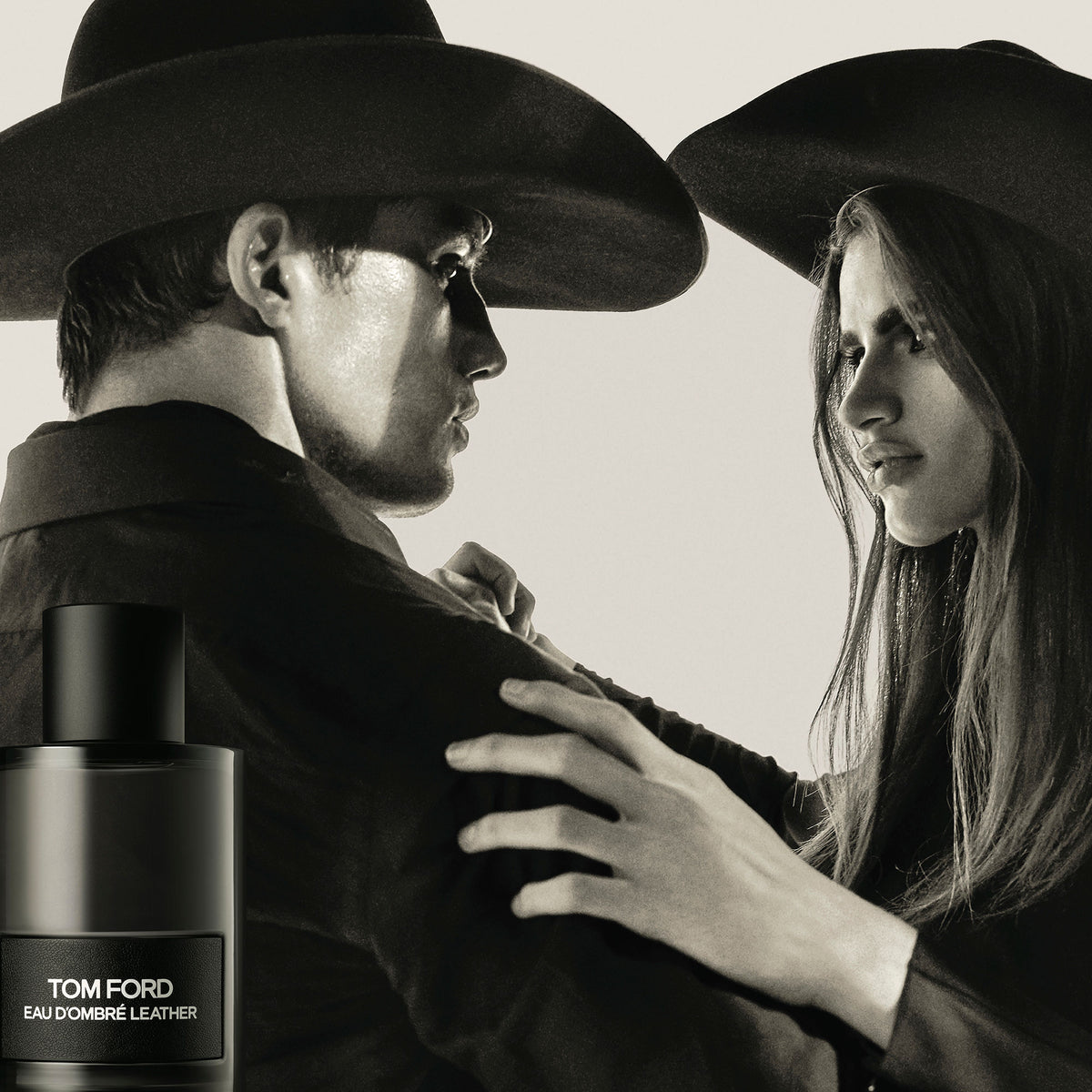 A man and woman wearing cowboy hats look at each other beside a bottle of Tom Ford Eau d’Ombre Leather Eau de Toilette, evoking notes of leather and amber.