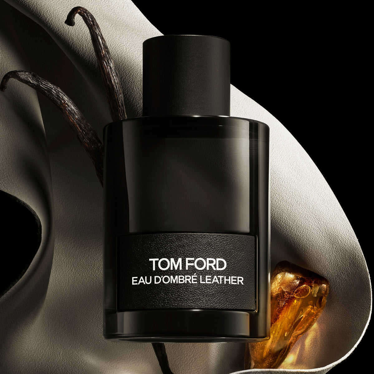 A black cylindrical bottle of Tom Ford Eau d’Ombre Leather Eau de Toilette rests on light fabric alongside aromatic vanilla beans and a smooth amber stone.