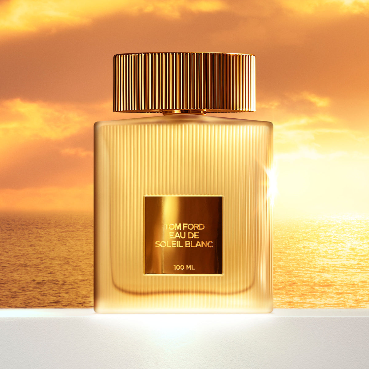 A rectangular Tom Ford Eau de Soleil Blanc Eau de Toilette bottle with a ribbed gold cap and glass body, centered against a sunset over the ocean, evoking floral warmth and sparkling citrus.