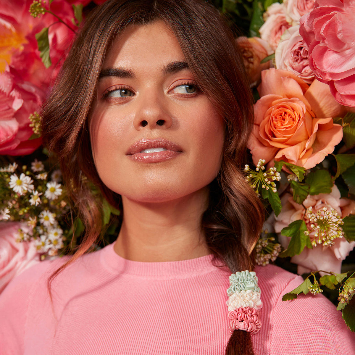 A woman wears three stacked pale green, white, and pink Slip Pure Silk Skinny Scrunchies around her braid in front of a backdrop of colorful flowers.