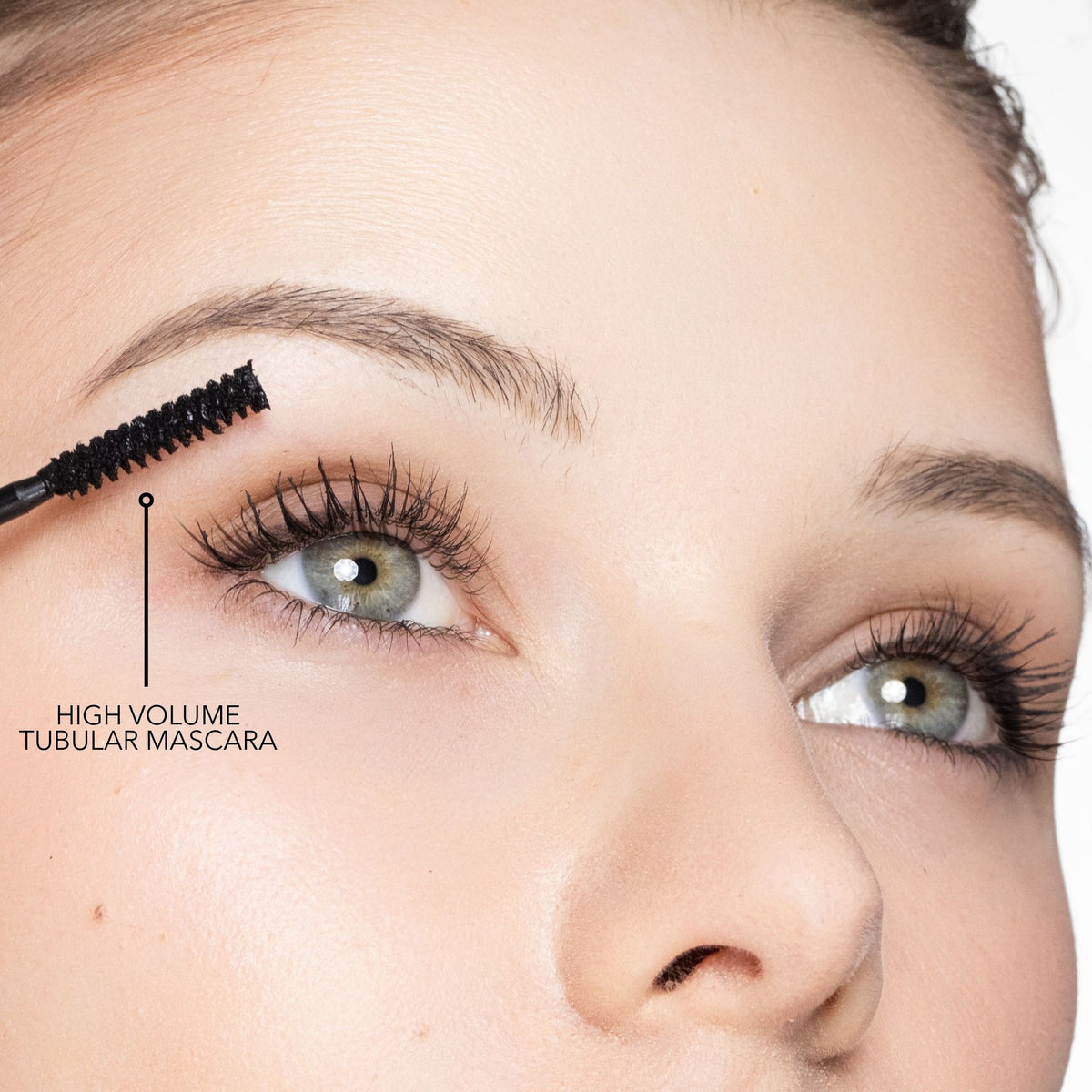 A close-up of an eye with a mascara wand applying The Trish McEvoy Tubular Lash Duo (Limited Edition) to volumize the lashes, with text reading HIGH VOLUME TUBULAR MASCARA.