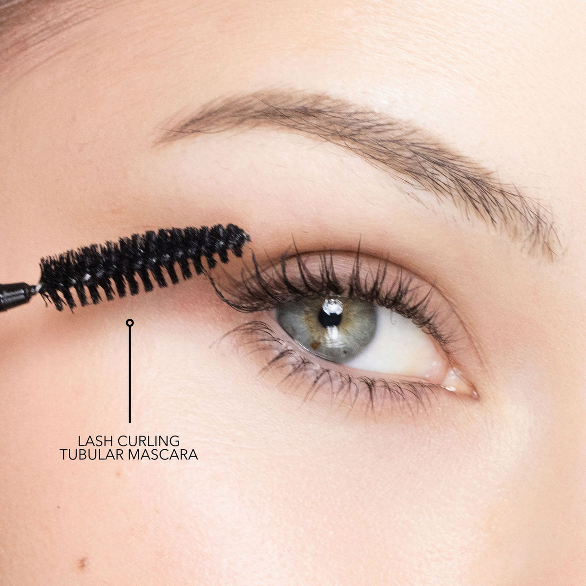 A Trish McEvoy Tubular Lash Duo mascara wand applies product to upper eyelashes, with a line pointing to the mascara to show how it curls lashes.