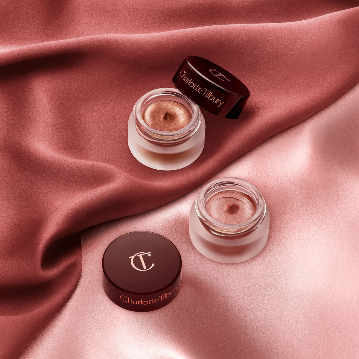 Two glass jars of Charlotte Tilbury Eyes To Mesmerise shimmering bronze cream eyeshadow with their lids placed beside them on draped rose-colored fabric.