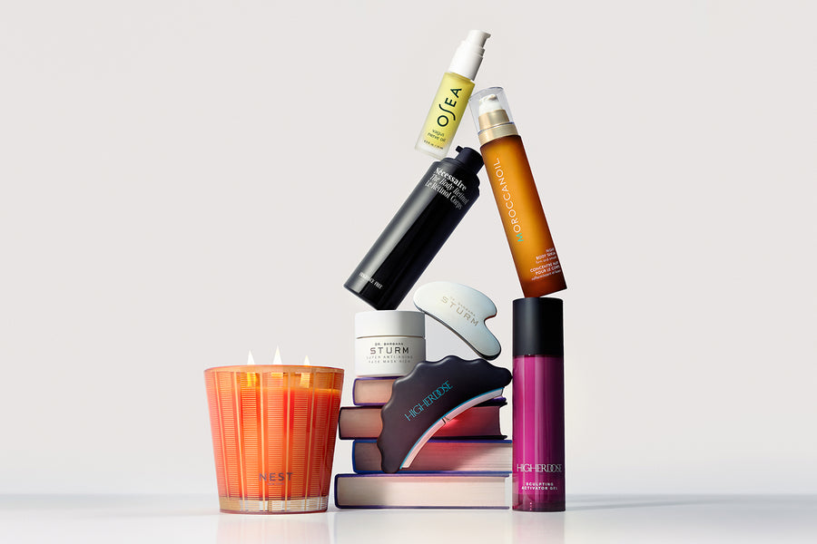 A styled arrangement of various beauty and fragrance products on a light gray background, including a lit orange candle, a black bottle, an amber spray bottle, a purple tube, and other cosmetic containers.

