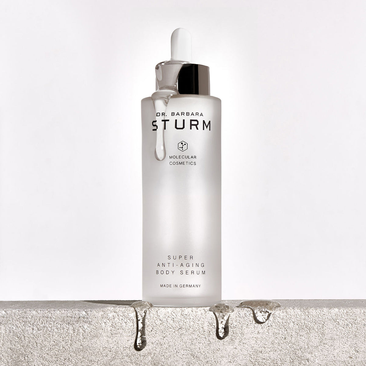A clear glass bottle of Dr. Barbara Sturm Super Anti-Aging Body Serum with a dropper dripping liquid, sitting on a rough stone surface with droplets of serum falling off the edge.