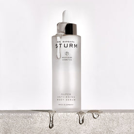 A clear glass bottle of Dr. Barbara Sturm Super Anti-Aging Body Serum with a dropper dripping liquid, sitting on a rough stone surface with droplets of serum falling off the edge.