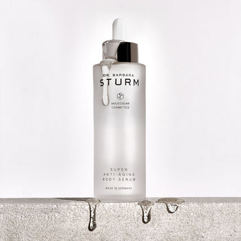 A clear glass bottle of Dr. Barbara Sturm Super Anti-Aging Body Serum with a dropper dripping liquid, sitting on a rough stone surface with droplets of serum falling off the edge.