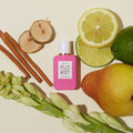 An Ellis Brooklyn Florist Eau de Parfum bright pink rectangular citrus floral bottle surrounded by a pear, lime slices, wood sticks, and tuberose stems.