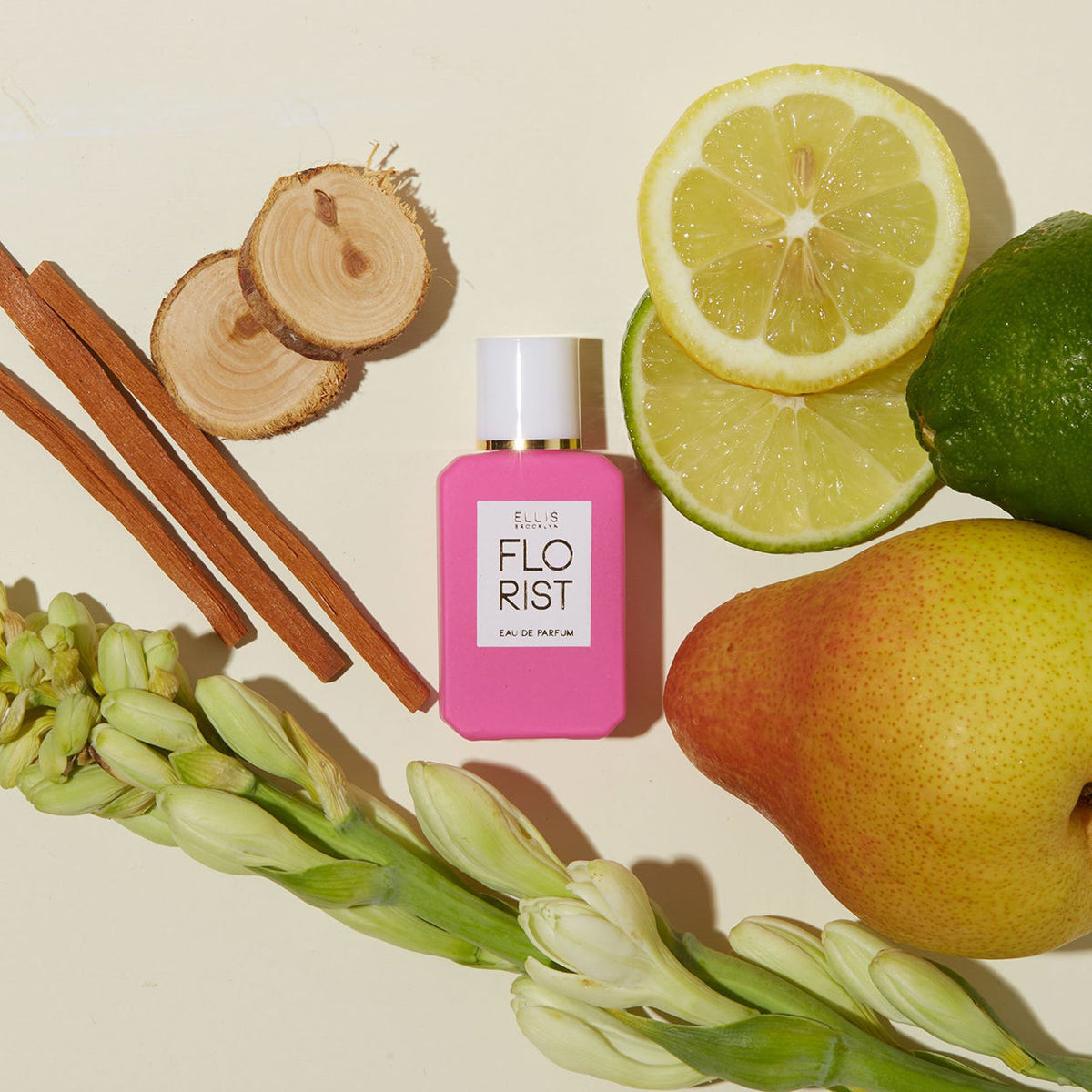 An Ellis Brooklyn Florist Eau de Parfum bright pink rectangular citrus floral bottle surrounded by a pear, lime slices, wood sticks, and tuberose stems.