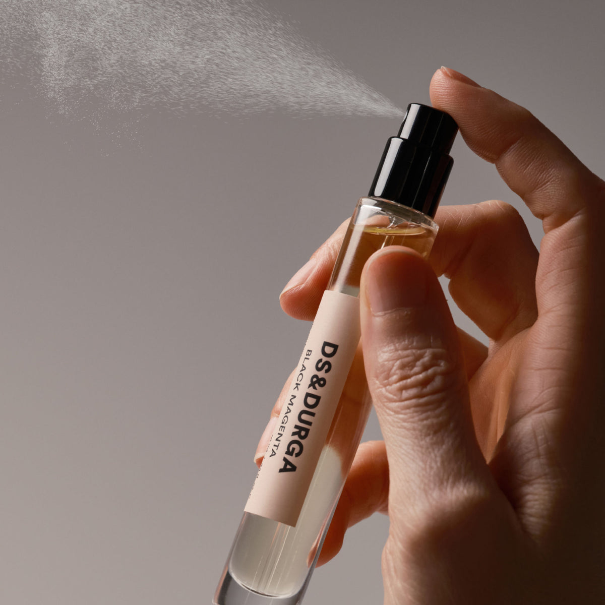 A hand presses the nozzle of a black amber Black Magenta Eau de Parfum bottle, releasing a fine mist into the night air.