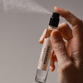 A hand presses the nozzle of a Rose Atlantic Eau de Parfum bottle, releasing a fine mist.