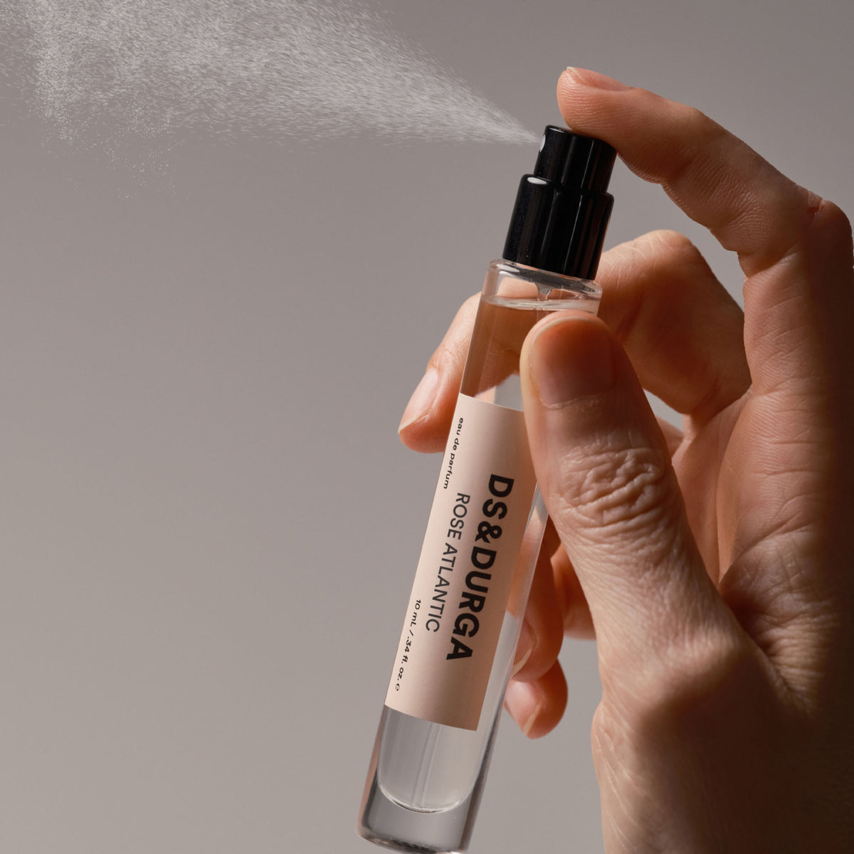 A hand presses the nozzle of a Rose Atlantic Eau de Parfum bottle, releasing a fine mist.