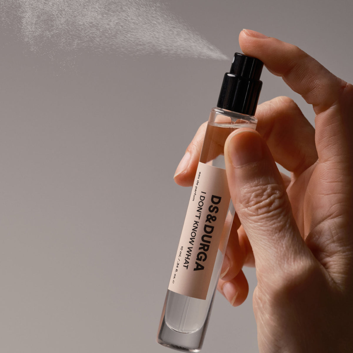 A hand sprays a small, clear glass vial of D.S. & Durga I Don’t Know What Eau de Parfum with a cream-colored label to reveal a sense of transparent radiance.