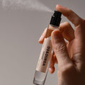 A hand presses the nozzle of a cylindrical glass bottle of Debaser Eau de Parfum, releasing a fine mist that hangs in the heavy August heat.
