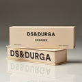 Two stacked, light tan rectangular D.S. & Durga Debaser Eau de Parfum boxes with the brand and product name in black text, sitting on a desk as indie rock plays over college radio.