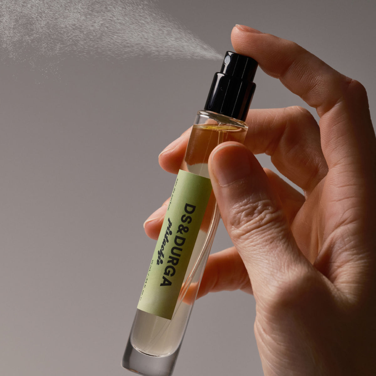 A hand holds a thin glass bottle of D.S. & Durga Pistachio Eau de Parfum with a green label as it releases a fine mist.