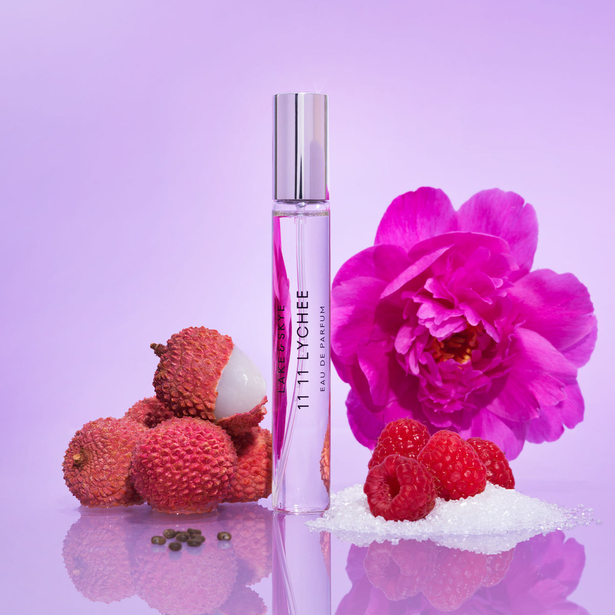 A Lake & Skye 11 11 Lychee Eau de Parfum glass rollerball bottle surrounded by lychee fruits, a large pink flower, and a small pile of raspberries and ambrette seed sugar.