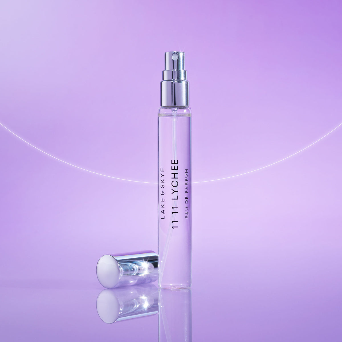 A tall, thin glass spray bottle of 11 11 Lychee Eau de Parfum with its silver cap resting on the surface next to it.