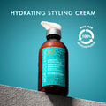 A pump bottle of Moroccanoil Hydrating Styling Cream sits on a stone ledge to help smooth fly-aways, featuring a circular icon indicating it is made from 100% recycled plastic.