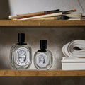 Two glass perfume bottles of different sizes sit on a wooden shelf next to a stack of L’Eau Papier Eau de Toilette, with paintbrushes resting on the shelf above.