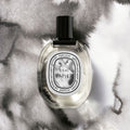 A Diptyque L’Eau Papier Eau de Toilette bottle with a black cap and an oval label set against a grey and white watercolor background, evoking a fresh, paper-inspired fragrance.