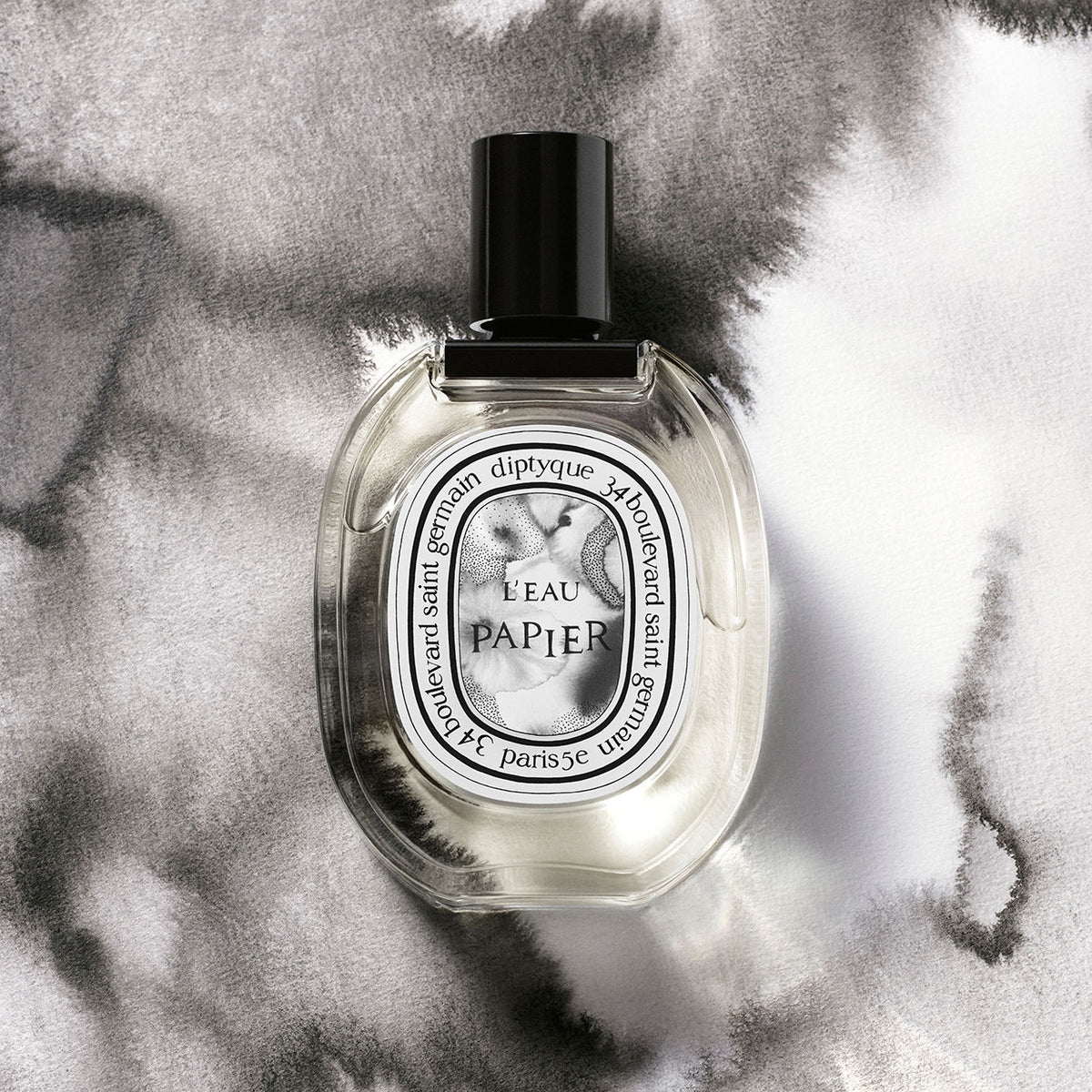 A Diptyque L’Eau Papier Eau de Toilette bottle with a black cap and an oval label set against a grey and white watercolor background, evoking a fresh, paper-inspired fragrance.