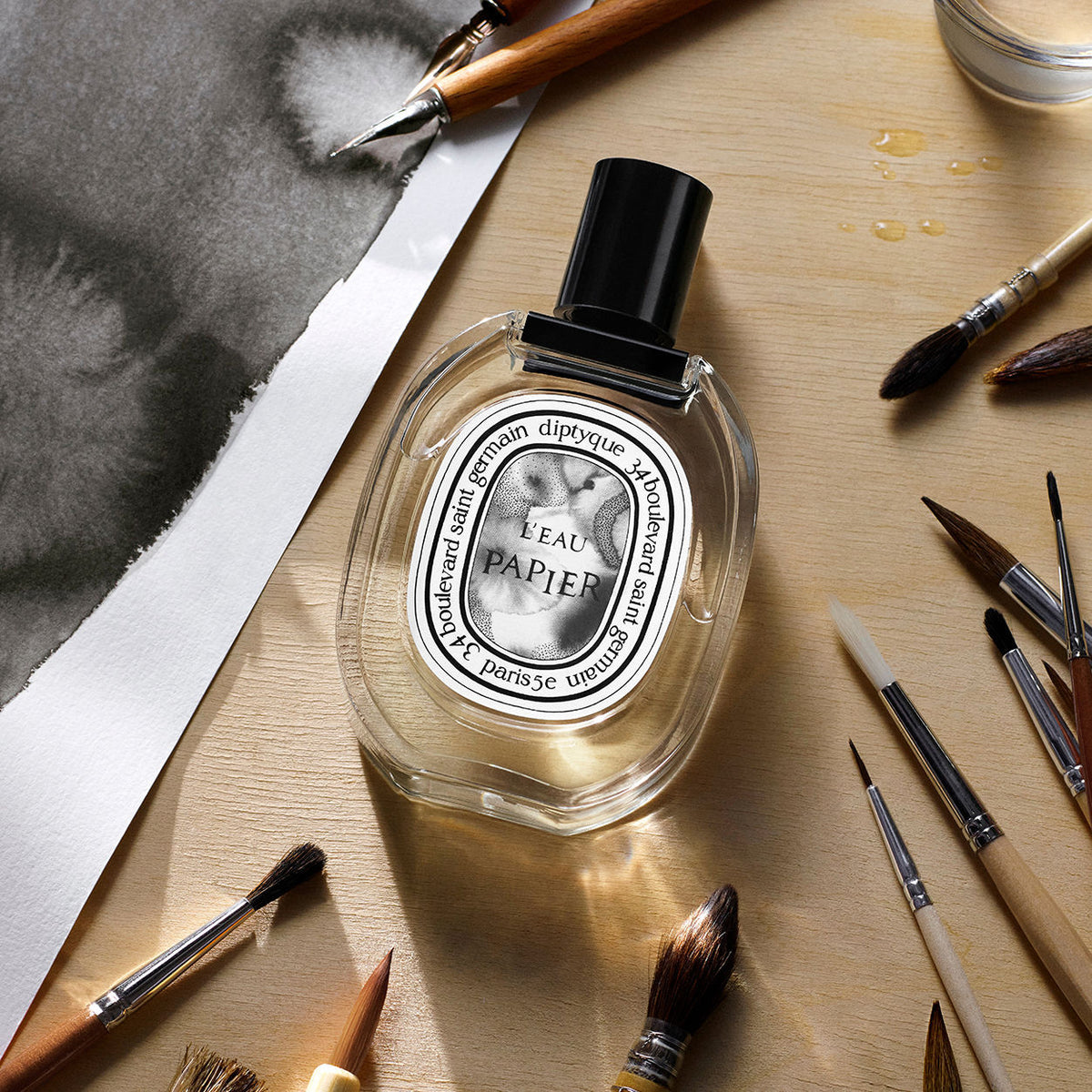 An oval Diptyque L’Eau Papier Eau de Toilette bottle with a black cap and label, surrounded by wooden paintbrushes and a piece of paper with a black ink wash, evoking a paper-inspired fragrance.