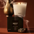 A Nest New York Moroccan Amber Candle in a glass jar sits atop its black box, next to a metallic vessel and a piece of raw amber.