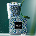 A patterned Nest New York Forest Rain Candle in a glass vessel sits atop its matching box, with a single green tulip and water droplets nearby, evoking the freshness of dewy fig leaves.
